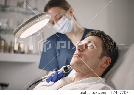 Young man laying in cosmetologist cabinet and has a cavitation procedure for face skin. Female cosmetologist wearing blue medical costume and face mask. Man wearing white bathrobe. 124377024