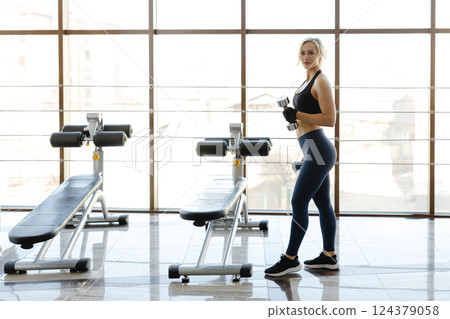 Determined sportswoman lifting dumbbells in a gym with natural light Determined sportswoman lifting dumbbells in a gym with natural light 124379058