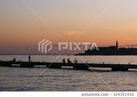 People on a pier at sunset by the Mediterranean Sea, silhouettes, old town in the background. 124381410