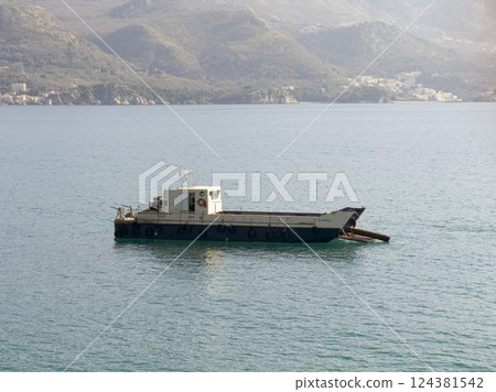 Old cargo barge floating on the calm blue Mediterranean Sea with mountains and a coastal town in the background 124381542
