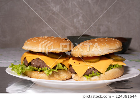 Two cheeseburgers with sesame buns, beef patties, melted cheddar, lettuce, pickles, and tomato sit on a white plate, ready for serving. Two cheeseburgers with sesame buns, beef patties, melted cheddar, lettuce, pickles, and tomato sit on a white plate, ready for serving. 124381644