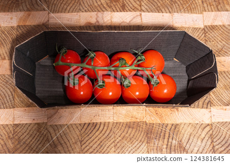 Red cherry tomatoes still attached to a vine, resting in a cardboard container on a patterned wooden cutting board. Red cherry tomatoes still attached to a vine, resting in a cardboard container on a patterned wooden cutting board. 124381645