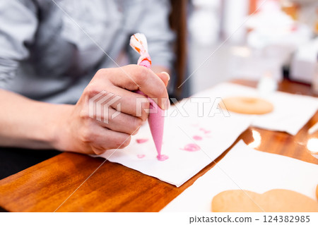 Adult female decorating cookies with pink icing using a piping bag in a kitchen setting 124382985