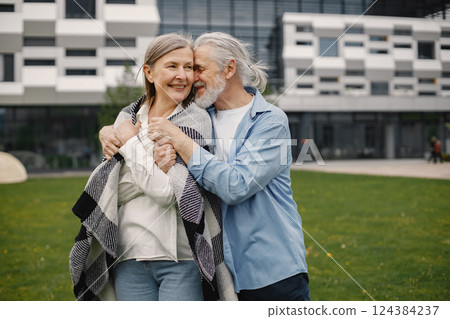 Caucasian elderly couple standing on a grass in summer. Man and woman hugging. Woman wearing white shirt and covering in plaid blanket. Caucasian elderly couple standing on a grass in summer. Man and woman hugging. Woman wearing white shirt and covering in plaid blanket. 124384237