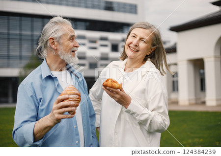 Caucasian elderly couple standing on a grass in summer. Man and woman holding a two croissants in hands. Woman wearing white shirt and man blue one. 124384239