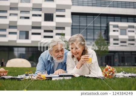 Caucasian elderly couple laying on a blanket on a grass in summer. Man and woman reading a book and drinking a juice. Woman wearing white shirt and man blue one. Caucasian elderly couple laying on a blanket on a grass in summer. Man and woman reading a book and drinking a juice. Woman wearing white shirt and man blue one. 124384249