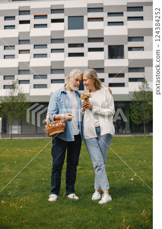 Caucasian elderly couple standing on a grass in summer. Man holding straw basket and woman flowers. Woman wearing white shirt and man blue one. 124384252