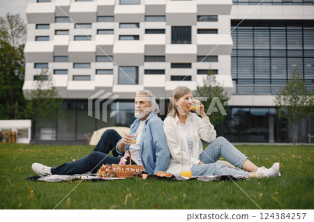 Beautiful caucasian elderly couple sitting on a grass in the park in summer. Man and woman sitting on blanket at the park drinking juice and sharing few precious memories. Woman wearing white shirt 124384257