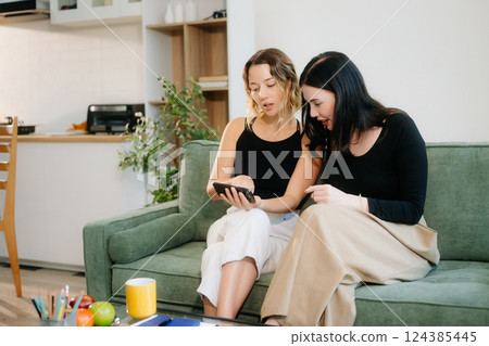 Two cheerful women sitting on a green couch, sharing ideas using a laptop and phone in a stylish home setup. 124385445