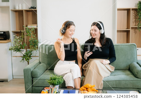 Two cheerful women sitting on a green couch, sharing ideas using a laptop and phone in a stylish home setup. Two cheerful women sitting on a green couch, sharing ideas using a laptop and phone in a stylish home setup. 124385458