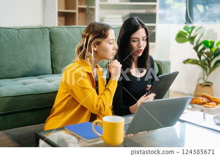 Two cheerful women sitting on a green couch, sharing ideas using a laptop and phone in a stylish home 124385671