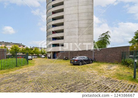 A sleek modern residential building towers over a structured parking area surrounded by greenery and a boundary wall, highlighting urban architecture. 124386667