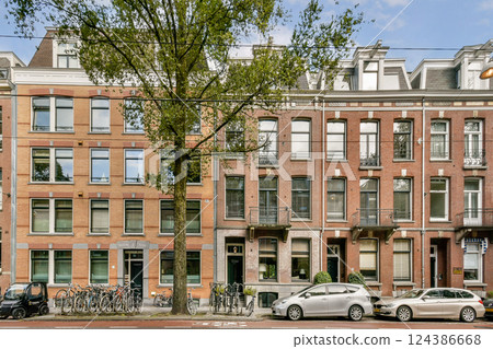 Brightly colored traditional buildings line a serene street, showcasing the unique architectural style of Amsterdam, complemented by bicycles and parked cars. 124386668