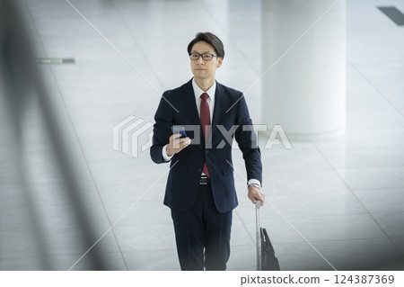 A businessman in his 40s walking with a smartphone and a suitcase 124387369