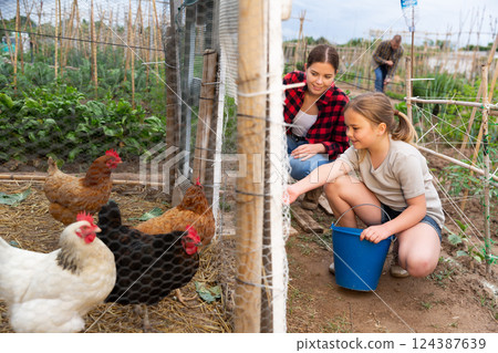 Mom and her daughter feed chickens in chicken coop in backyard of country house Mom and her daughter feed chickens in chicken coop in backyard of country house 124387639