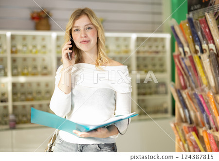 Girl employee of perfume shop talks on mobile phone, holding folder with documents in her hands 124387815