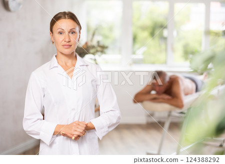Positive middle-aged masseuse posing against background of therapy room 124388292