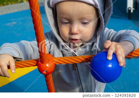 Toddler girl with excited and amused expression enjoys crawling in grey slide tube on playground. Child laughs and smiles playing in city park 124391620