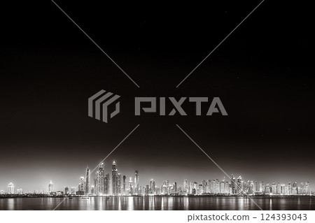Monochrome city skyline with tall skyscrapers reflecting on calm water under a cloudy sky, Dubai Marina bay UAE 124393043