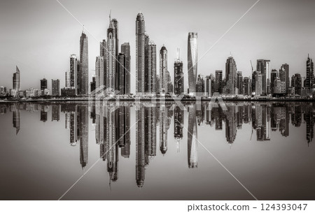 Monochrome city skyline with tall skyscrapers reflecting on calm water under a cloudy sky, Dubai Marina bay UAE 124393047