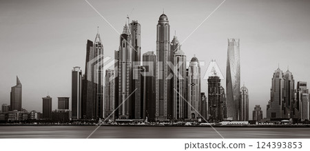 Monochrome city skyline with tall skyscrapers reflecting on calm water under a cloudy sky, Dubai Marina bay UAE 124393853