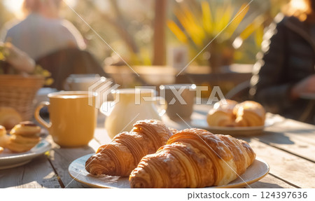 Fresh croissants on breakfast table in warm morning sunlight, people gathered in the background, creating cozy, inviting atmosphere for brunch or coffee or tea 124397636