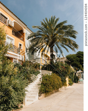 White staircase leading to colorful mediterranean houses with balconies and tall palm tree surrounded by lush vegetation in sunny summer day. Tall palm tree framing lush greenery under bright sunlight White staircase leading to colorful mediterranean houses with balconies and tall palm tree surrounded by lush vegetation in sunny summer day. Tall palm tree framing lush greenery under bright sunlight 124404094