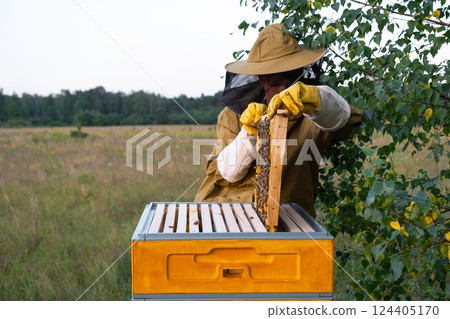 A beekeeper, a woman in a protective suit against bee stings, holds a frame with honey from a bee hive in her hands. Beekeeping, care of the hive 124405170
