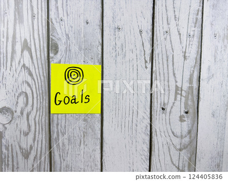 Sticky note showcasing the word Goals accompanied by target symbol, set against textured wooden background, symbolizing the importance of setting and achieving personal aspirations 124405836