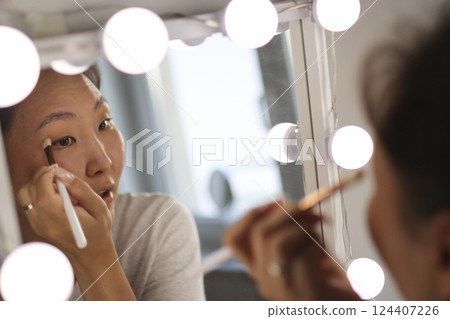 Asian woman applying makeup in front of mirror with lights 124407226