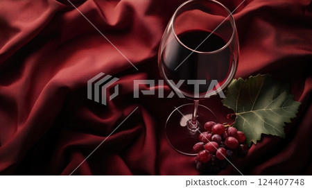 a glass of red wine placed on luxurious deep red fabric, accompanied by a cluster of grapes and a green vine leaf 124407748