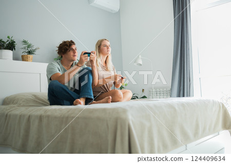 Young couple in love playing video games while lying on the bed in the bedroom, having fun and enjoying each other on their weekends. Technologies and gaming concept Young couple in love playing video games while lying on the bed in the bedroom, having fun and enjoying each other on their weekends. Technologies and gaming concept 124409634