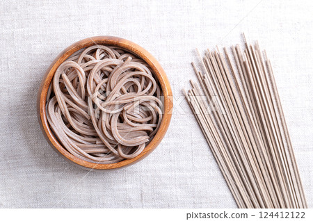 Chilled and boiled soba noodles in a wooden bowl on linen fabric. Thin Japanese noodles, made primarily from buckwheat flour, with wheat flour mixed in. With dried noodles placed on the right side. Chilled and boiled soba noodles in a wooden bowl on linen fabric. Thin Japanese noodles, made primarily from buckwheat flour, with wheat flour mixed in. With dried noodles placed on the right side. 124412122
