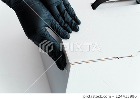 Close-up View of Hand Wearing Black Gloves Handling White Cardboard Box 124413990
