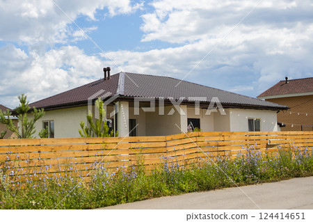 One-story house behind wooden fence and blooming wildflowers. Suburban privacy and residential space integration with natural elements. One-story house behind wooden fence and blooming wildflowers. Suburban privacy and residential space integration with natural elements. 124414651