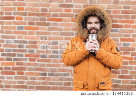 Man with cup looks away on winter street near brick wall with copy space. Seasonal comfort, morning routine, and emotional reflection outdoors. Man with cup looks away on winter street near brick wall with copy space. Seasonal comfort, morning routine, and emotional reflection outdoors. 124415398