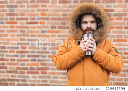 Man with cup looks away on winter street near brick wall with copy space. Seasonal comfort, morning routine, and emotional reflection outdoors. 124415399