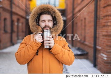 Man in winter parka holds thermos with closed eyes on snowy urban street. Warmth, solitude, and comfort in cold weather. 124415403