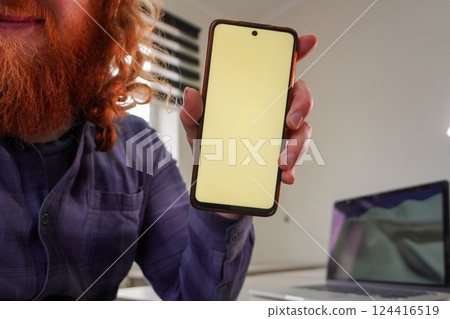 A cheerful man with a red beard showcases a blank smartphone screen, emphasizing modern technology and personal connection in a bright, minimalistic workspace. A cheerful man with a red beard showcases a blank smartphone screen, emphasizing modern technology and personal connection in a bright, minimalistic workspace. 124416519