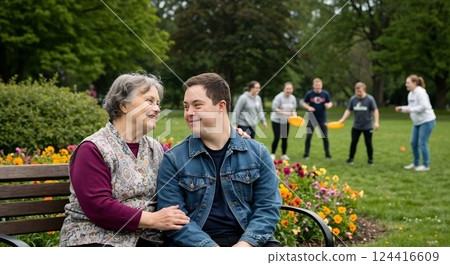 Shared Moment in the Park: A heartwarming scene unfolds in a park as a senior woman gazes lovingly at a young man, while a group plays in the background. 124416609