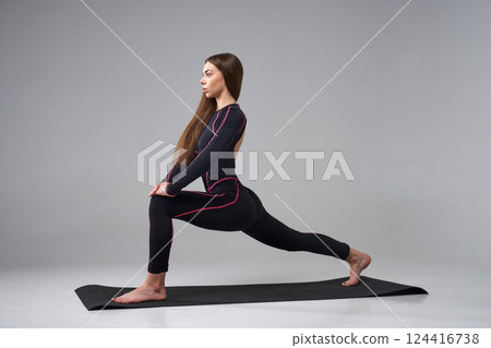 Slim woman in black bodysuit exercising on gym mat. Side view of calm Caucasian girl with long hair, practicing yoga, isolated on grey studio background. Concept of yoga, sport, healthy lifestyle. 124416738
