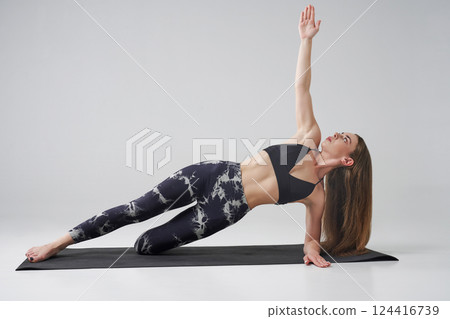 Calm female in activewear doing variation of side plank with bent leg indoors. Side view of barefoot sportswoman practicing side plank yoga position, isolated on grey background. Yoga concept. Calm female in activewear doing variation of side plank with bent leg indoors. Side view of barefoot sportswoman practicing side plank yoga position, isolated on grey background. Yoga concept. 124416739