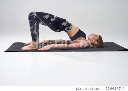 Mindful slim woman in leggings doing bridge position of yoga. Side view of barefoot female in activewear in Setu Bandhasana yoga pose on mat, during training, isolated on grey. Concept of yoga. Mindful slim woman in leggings doing bridge position of yoga. Side view of barefoot female in activewear in Setu Bandhasana yoga pose on mat, during training, isolated on grey. Concept of yoga. 124416741