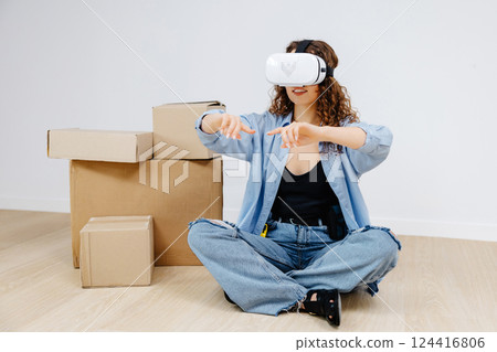 Young woman in VR glasses using modern technology creating room design, moving hands in air, sitting in new apartment during renovation. Virtual reality and repair concept. 124416806