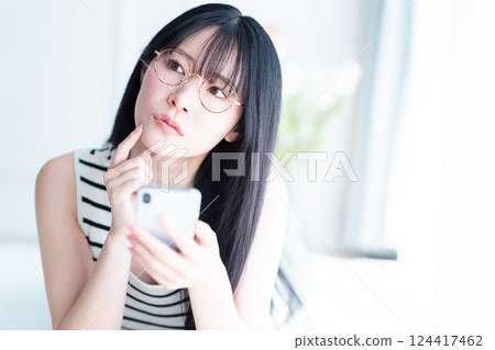 A young woman wearing glasses holding a smartphone and wondering what to buy A young woman wearing glasses holding a smartphone and wondering what to buy 124417462