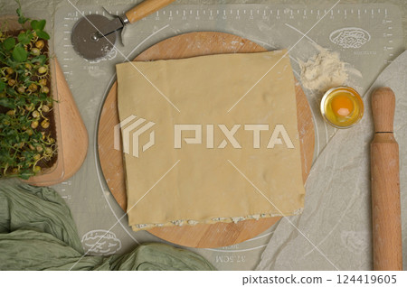 Top view square sheet of dough on wooden board. Rolling pin, cutter, egg, green towel, microgreens sprouts on kitchen table. Home cooking baking puffs 124419605