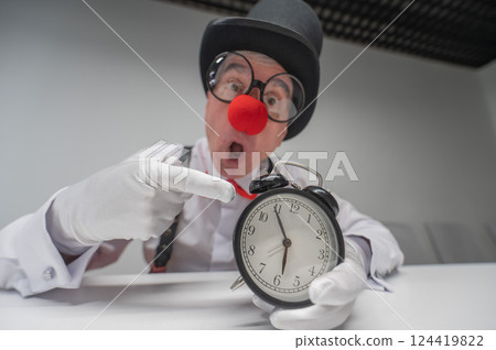 An elderly man in a clown costume points at an alarm clock.  124419822