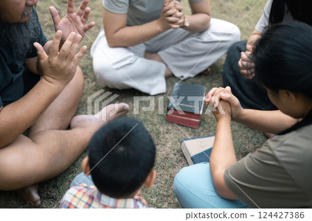 Young woman held her mother's hand in prayer, surrounded by their outdoor community, where love and family life flourished through shared moments of prayer and support. Group christian pray concept. 124427386