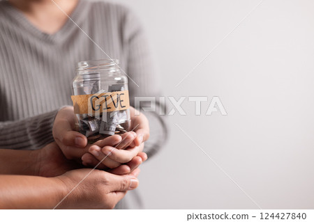 Woman held jar filled with cash with words in bold lettering urging people to donate money, extending her hand in gesture of giving part of her business's charitable efforts. Woman held jar filled with cash with words in bold lettering urging people to donate money, extending her hand in gesture of giving part of her business's charitable efforts. 124427840