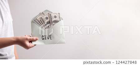 Woman's hand holding dollar bills in envelope with the letters word give, extended her hand to donate cash, promoting the spirit of giving in her business. banner with copy space. Woman's hand holding dollar bills in envelope with the letters word give, extended her hand to donate cash, promoting the spirit of giving in her business. banner with copy space. 124427848